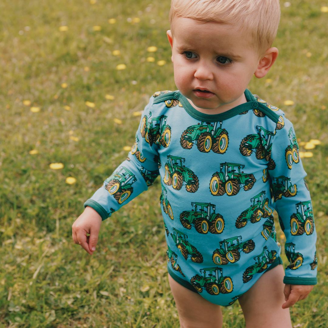 Child wearing a blue onesie with green tractor patterns outdoors on grass.