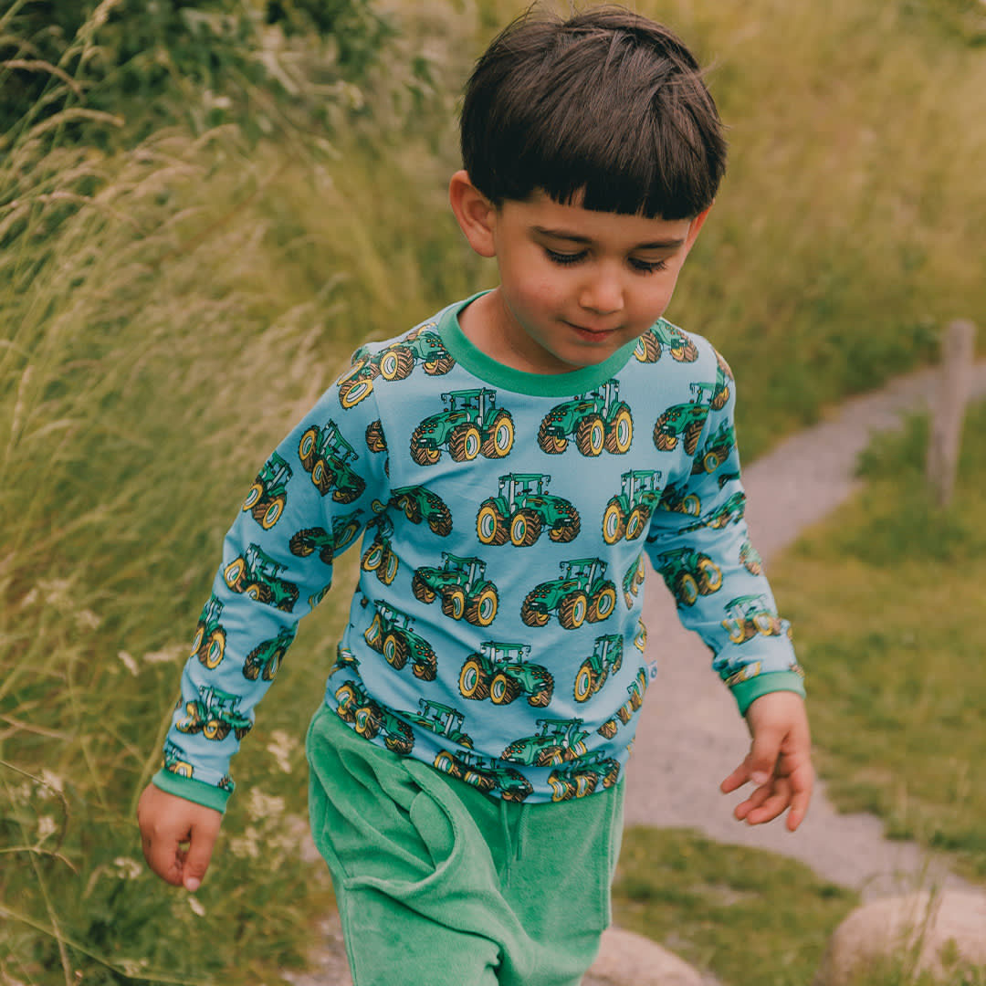 Child wearing a blue shirt with green tractor patterns and green pants, standing in a grassy field.