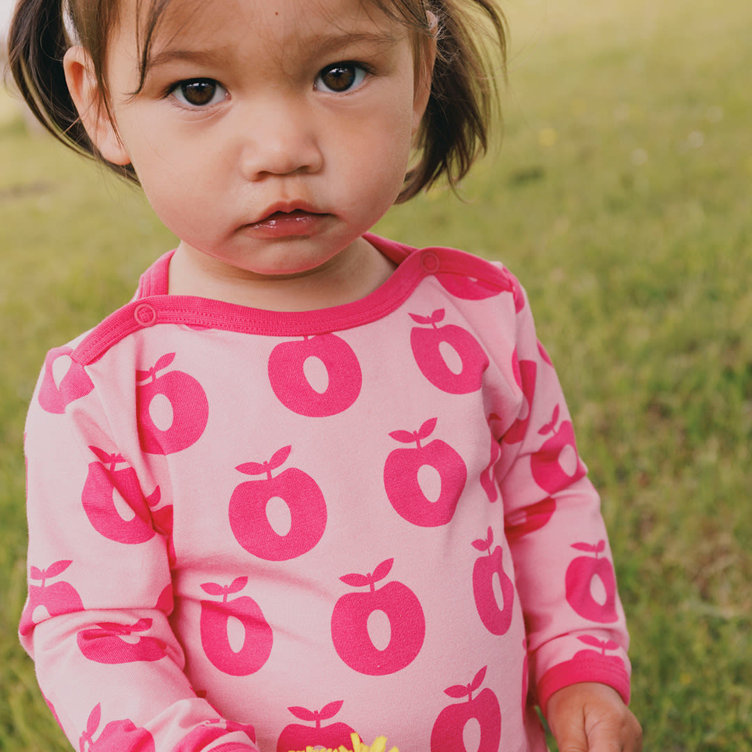 Child wearing a pink onesie with red apple patterns in a grassy field